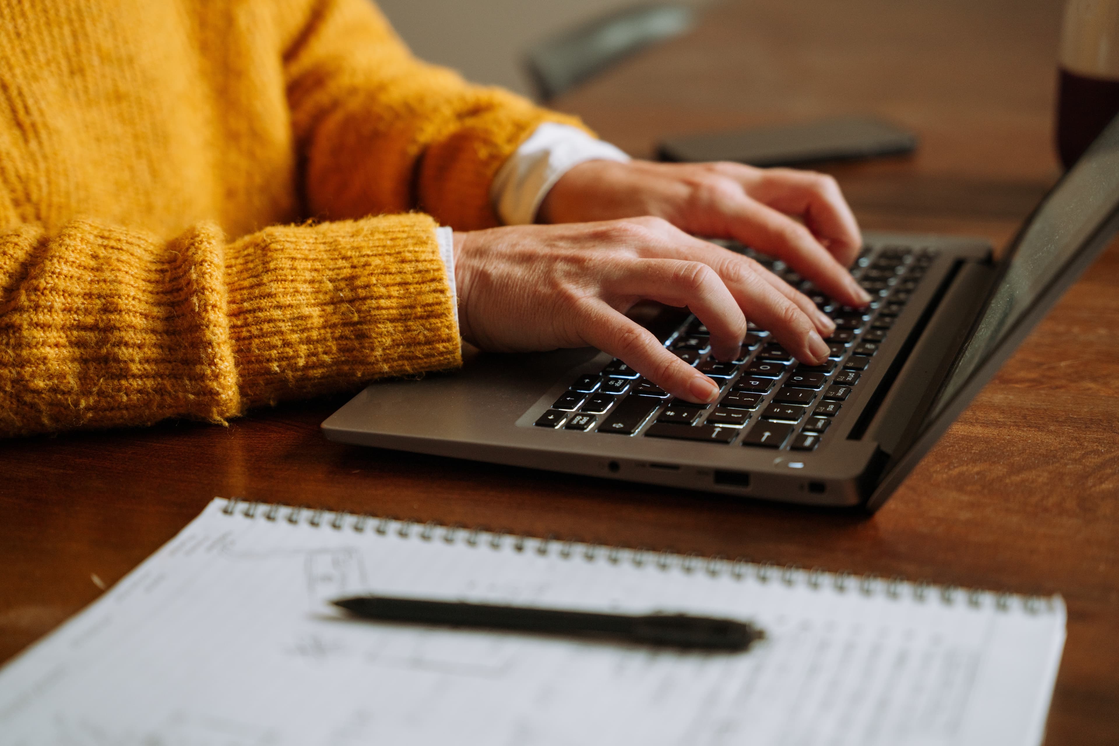 Person wearing a yellow sweater typing on a laptop at a wooden table, with a pen and notebook containing handwritten notes in the foreground.